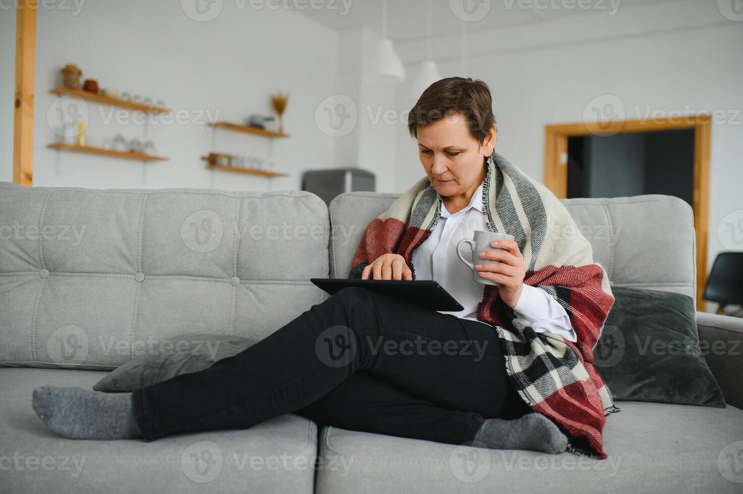 Peaceful satisfied mature woman wrapped warm blanket reading book, favorite literature and drinking tea or coffee, happy middle aged senior female relaxing, enjoying free time, weekend at home photo