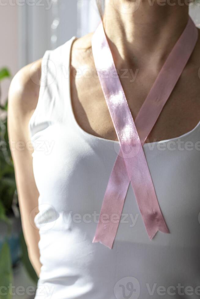 Shot of the woman in the white top against the white wall, with pink ribbon on her neck as a symbol of breast cancer awareness. Concept photo