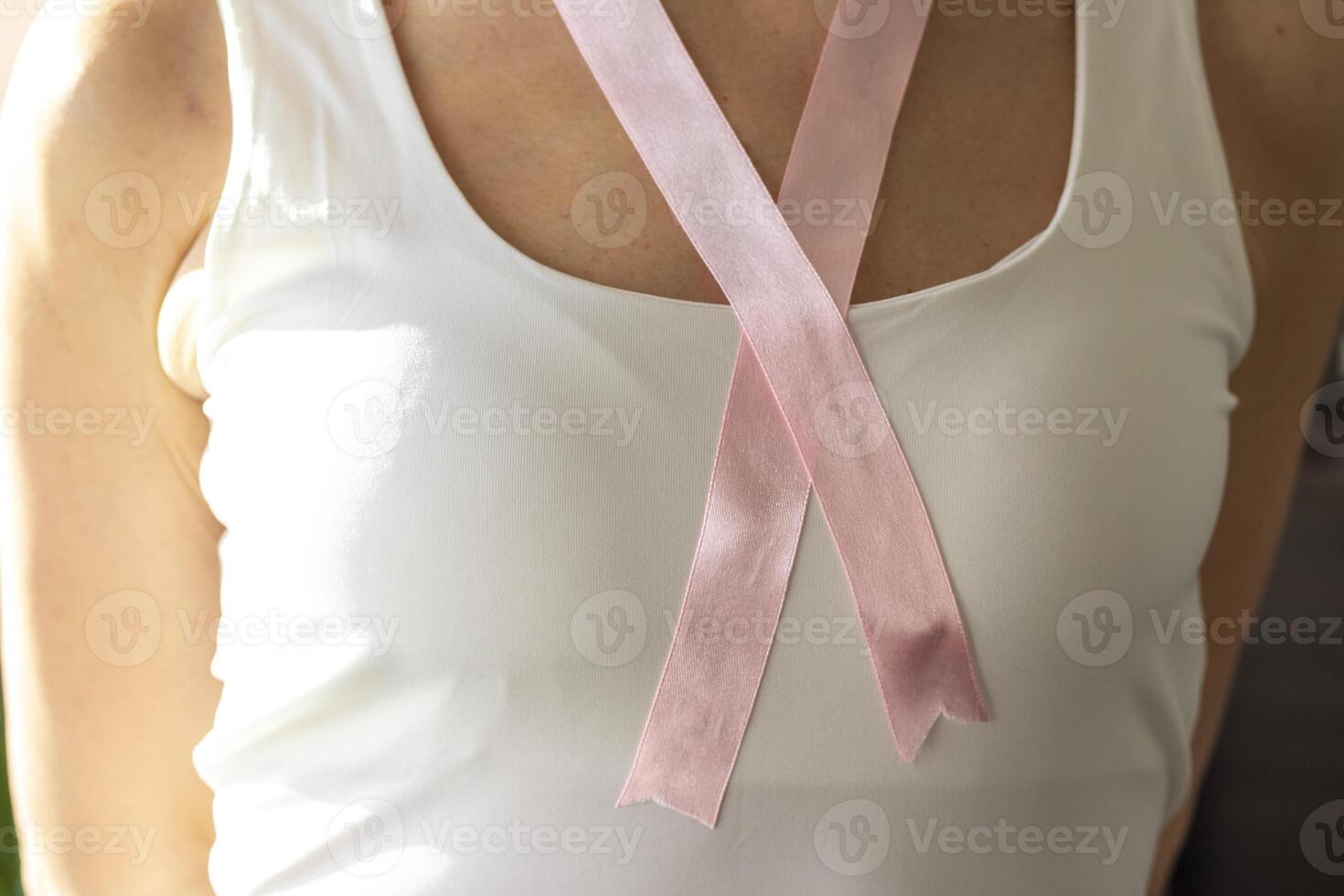 Shot of the woman in the white top against the white wall, with pink ribbon on her neck as a symbol of breast cancer awareness. Concept photo