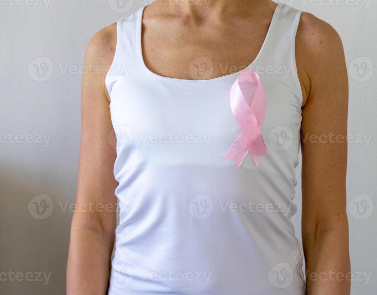 Shot of the woman against the white wall in the white top with pink ribbon, as a symbol of a breast cancer awareness. Concept photo