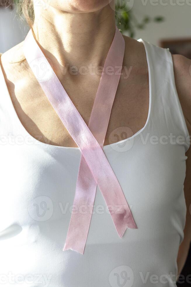 Shot of the woman in the white top against the white wall, with pink ribbon on her neck as a symbol of breast cancer awareness. Concept photo