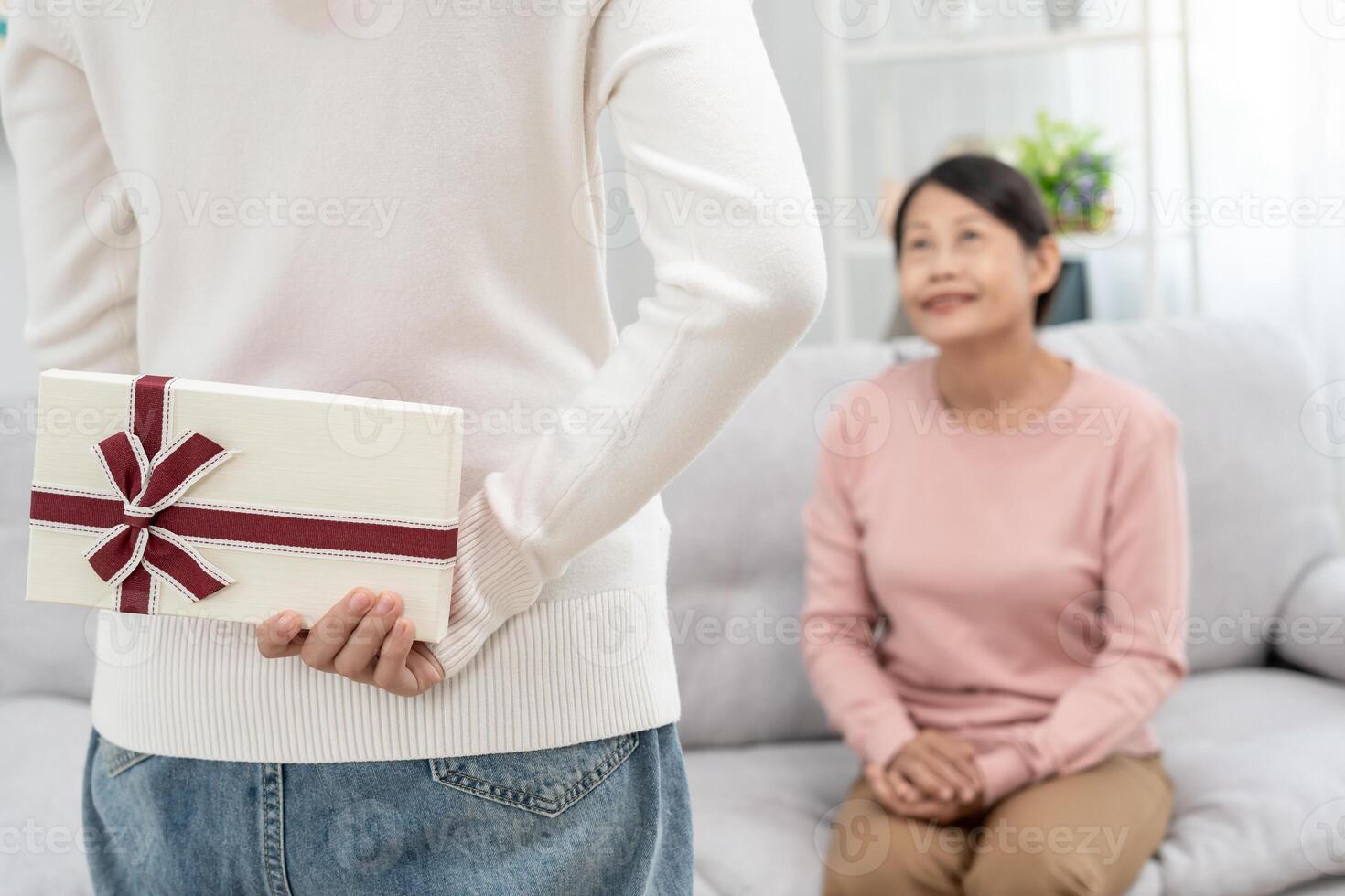 teen girls are hiding gift boxes for their beautiful moms behind their backs while mom is sitting on the sofa at home. celebrating special occasion together at home, happy birthday to mom photo