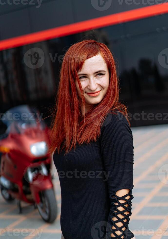 un elegante pelirrojo posando siguiente a un pulcro motocicleta. un mujer con rojo pelo en pie siguiente a un motocicleta foto