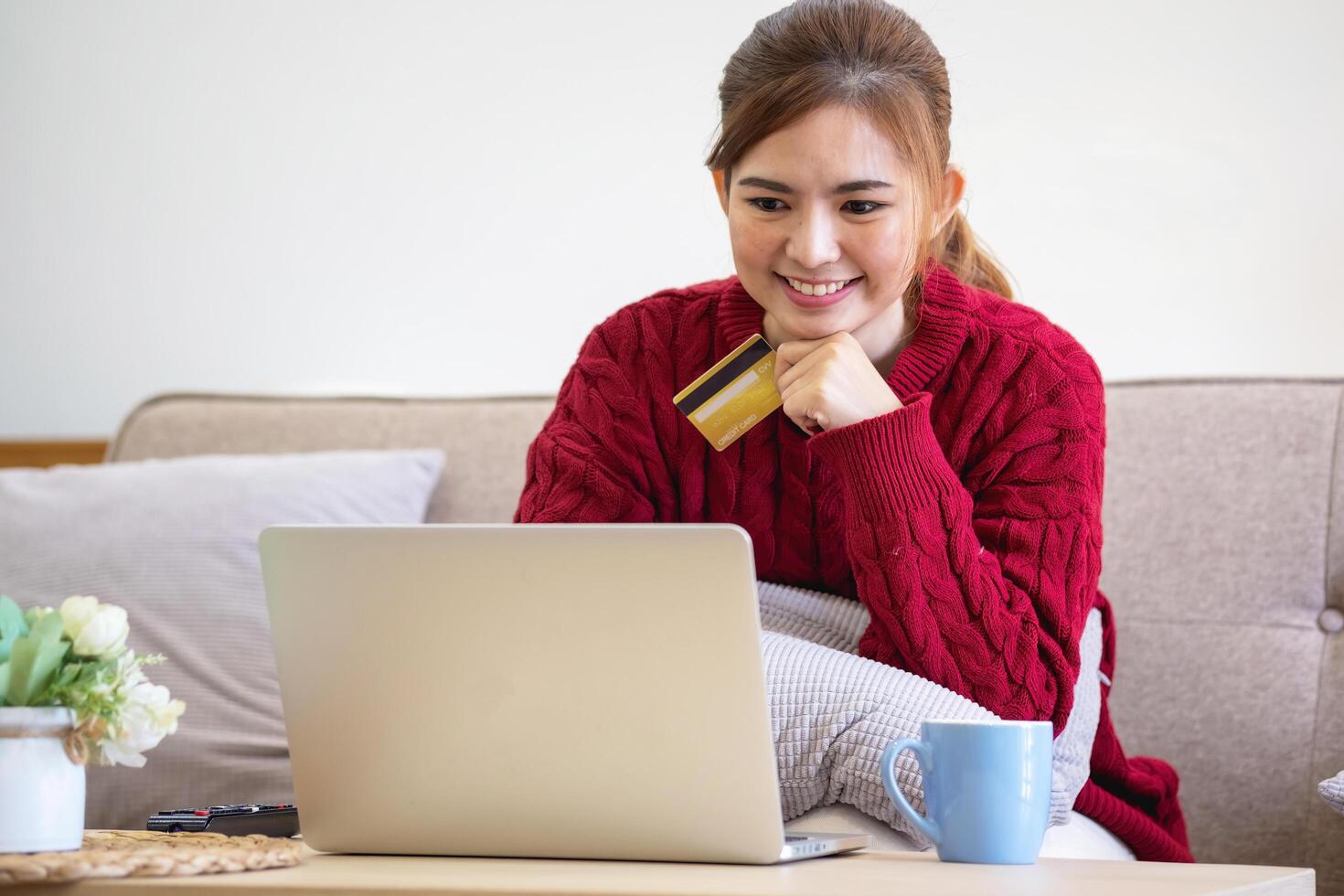 un joven asiático mujer con un contento sonrisa sostiene un crédito tarjeta y usos un teléfono inteligente a tienda en línea en línea pago concepto. foto