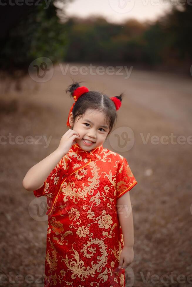 alegre joven niña con adorable rojo pelo bollos vistiendo un vibrante tradicional chino cheongsam vestir al aire libre en el jardín parque. chino día concepto. foto