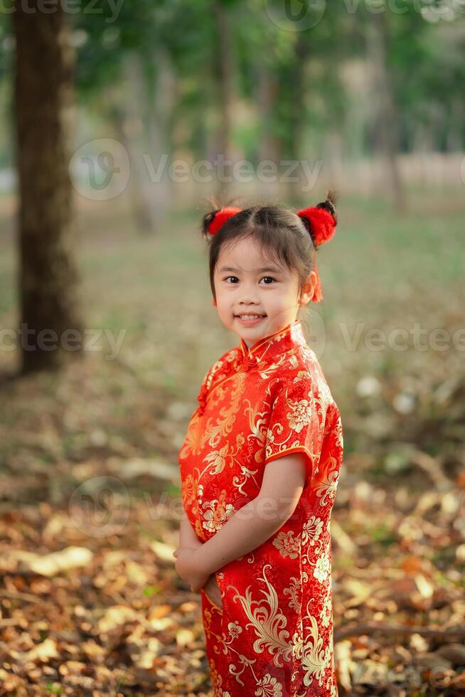 Cheerful young girl with adorable red hair buns wearing a vibrant traditional Chinese cheongsam dress outdoors in the garden park. Chinese day concept. photo