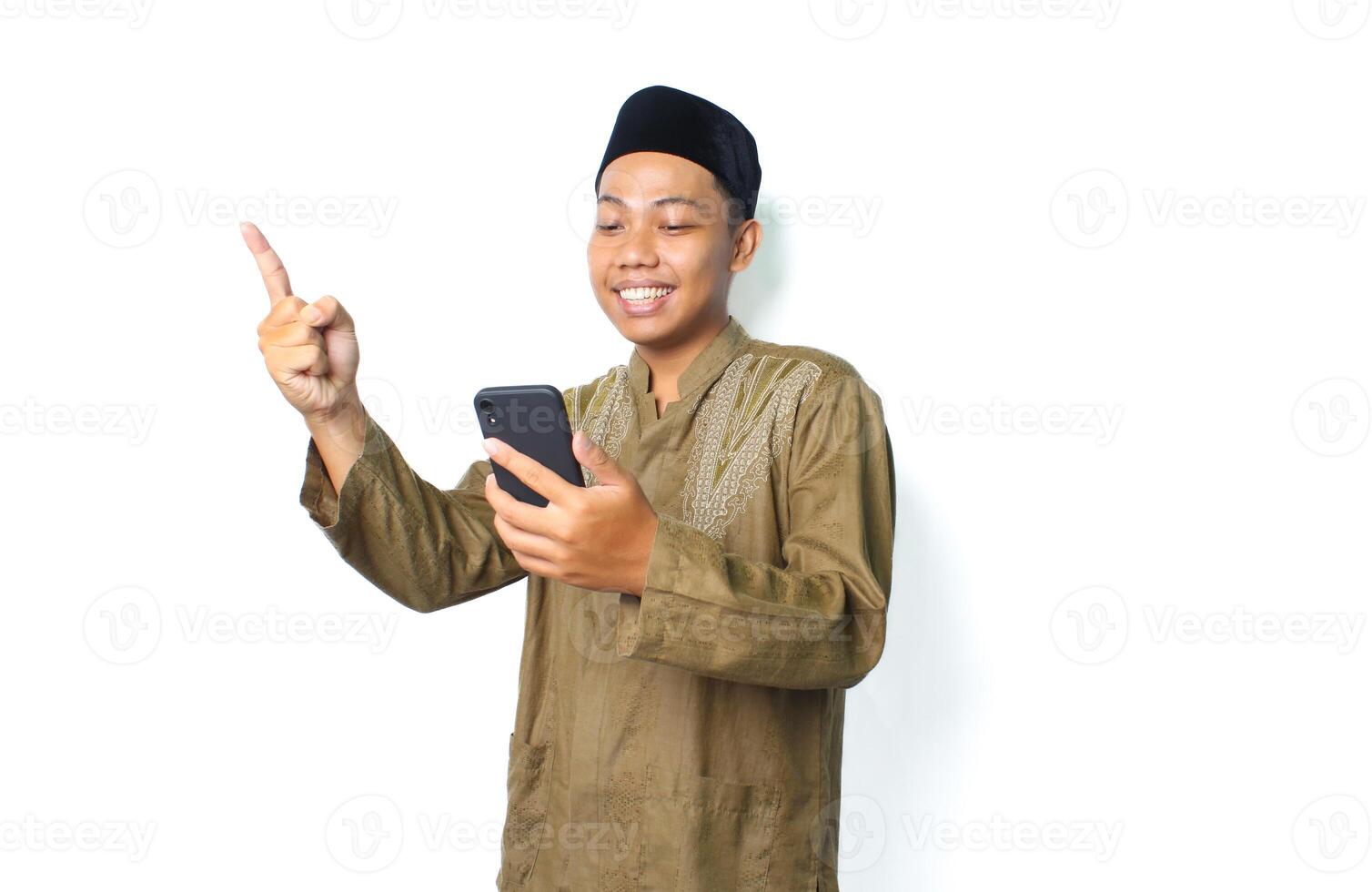 excited asian muslim male holding mobile phone and pointing to beside isolated on white background photo