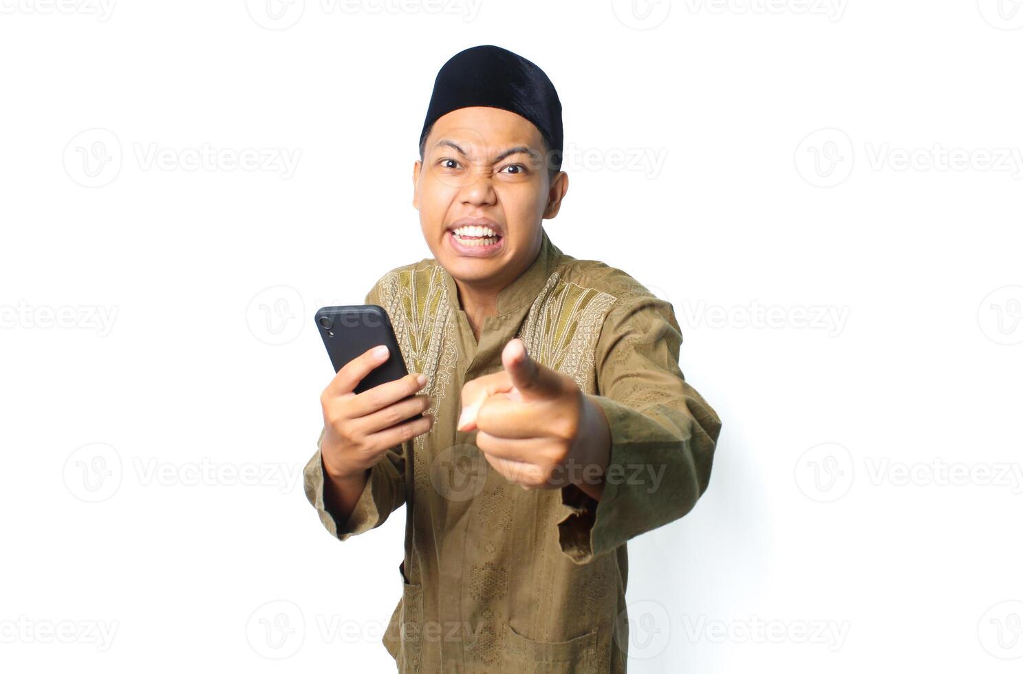 emotional asian muslim man pointing at camera with holding mobile phone isolated on white background photo