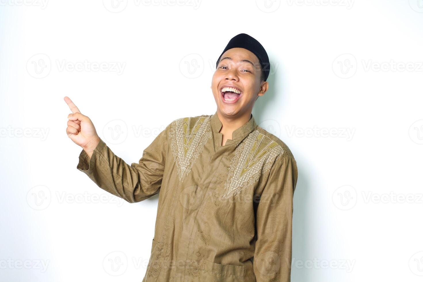 ecstatic asian muslim man wearing islamic dress pointing to empty side with laugh isolated on white background photo