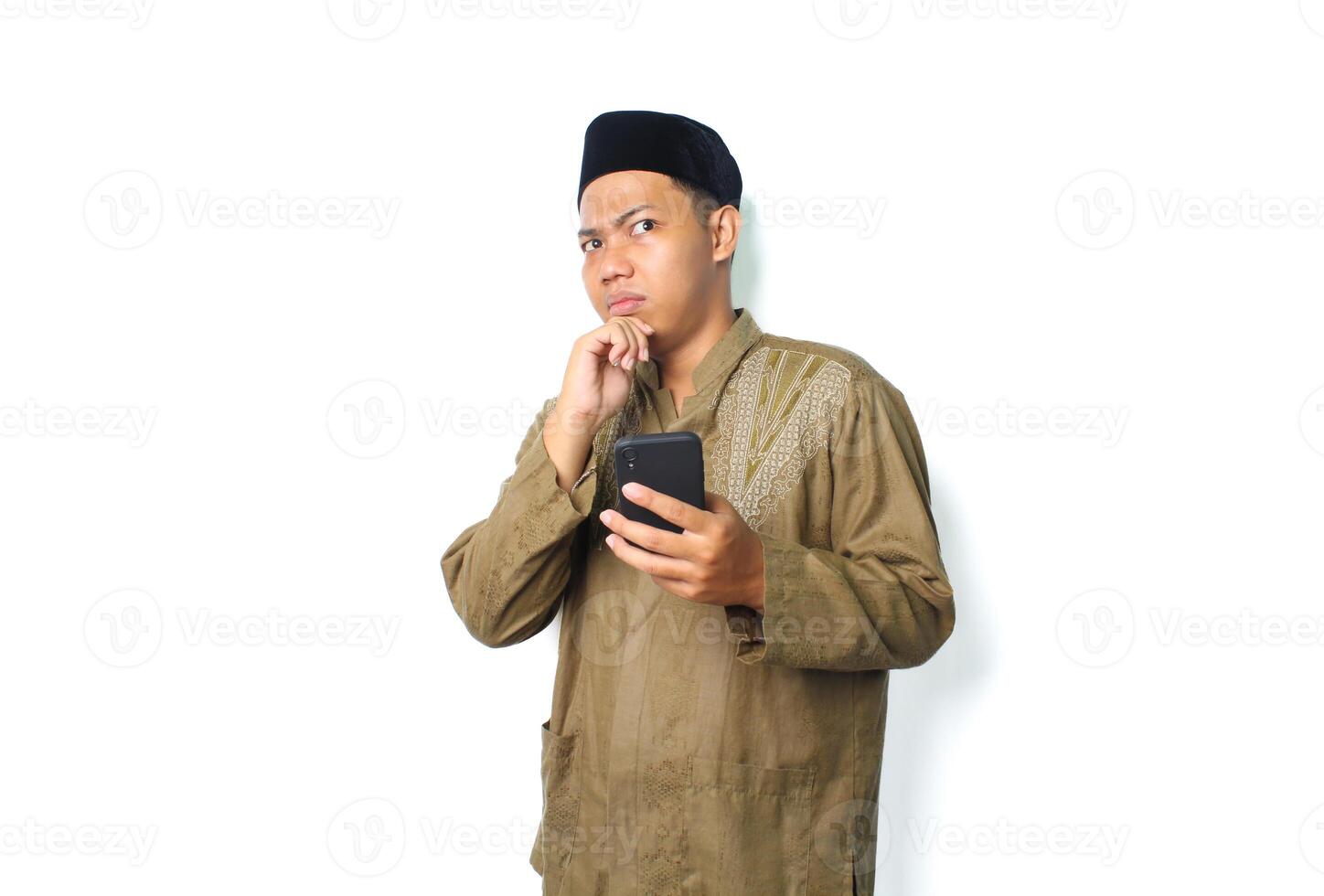 thoughtful asian muslim man holding mobile phone with thouching his chin looking away isolated on white background photo