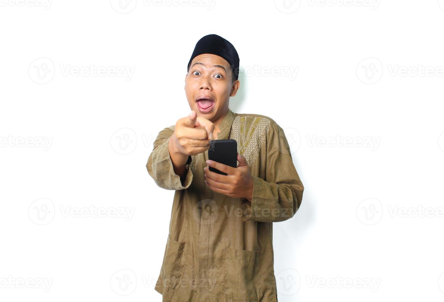 surprised asian muslim man wearing islamic clothes pointing at camera with holding mobile phone isolated on whtie background photo