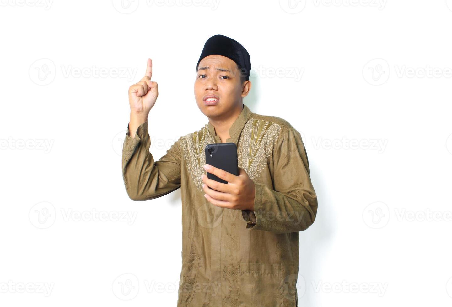 anxious asian muslim man holding mobile phone with pointing to above show worry expression isolated on white background photo