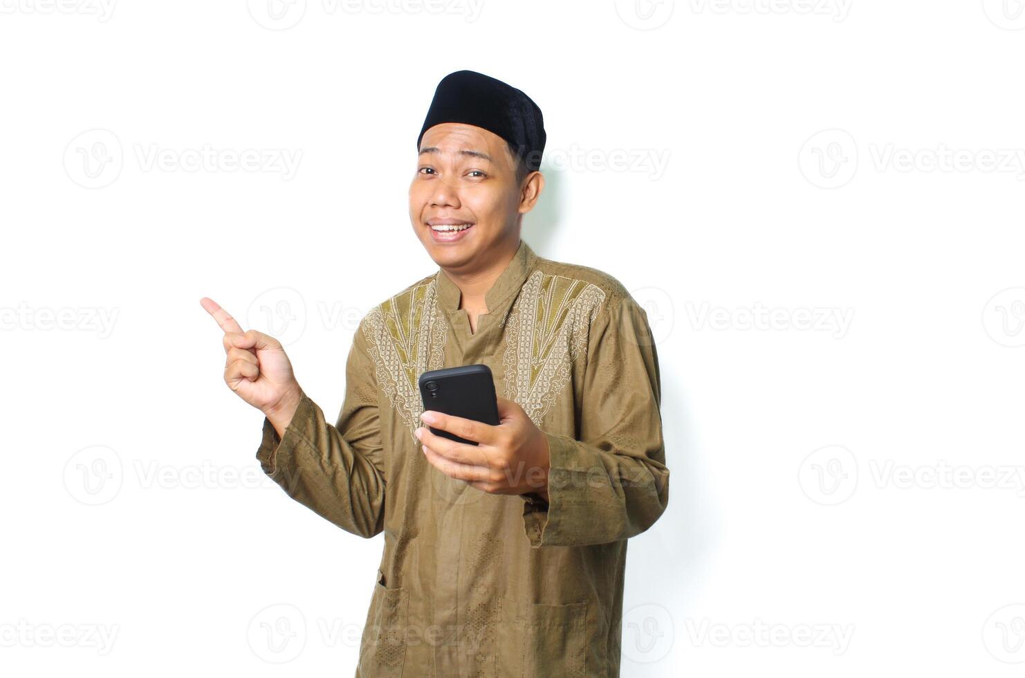 funny asian muslim man pointing to side with holding smartphone isolated on white background photo