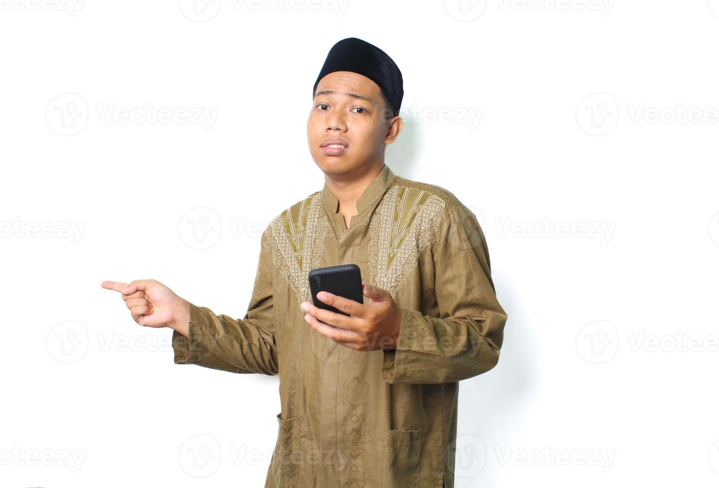 anxious asian muslim man holding mobile phone with pointing to beside show worry expression isolated on white background photo