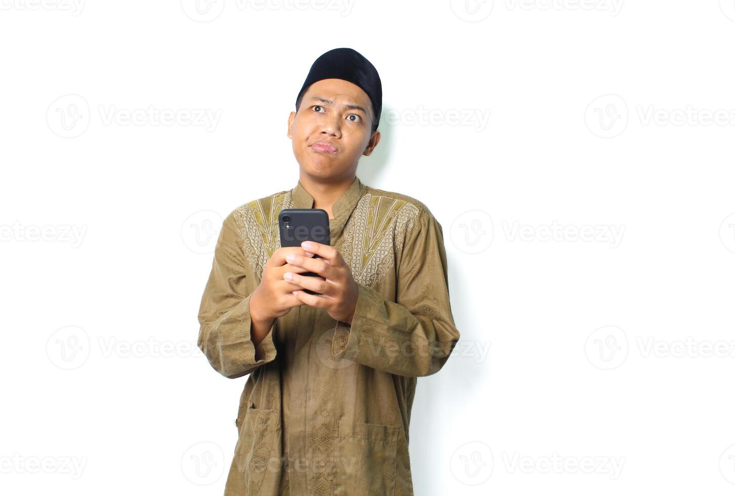 confused asian muslim man holding mobile phone looking away remembering memory isolated on white background photo