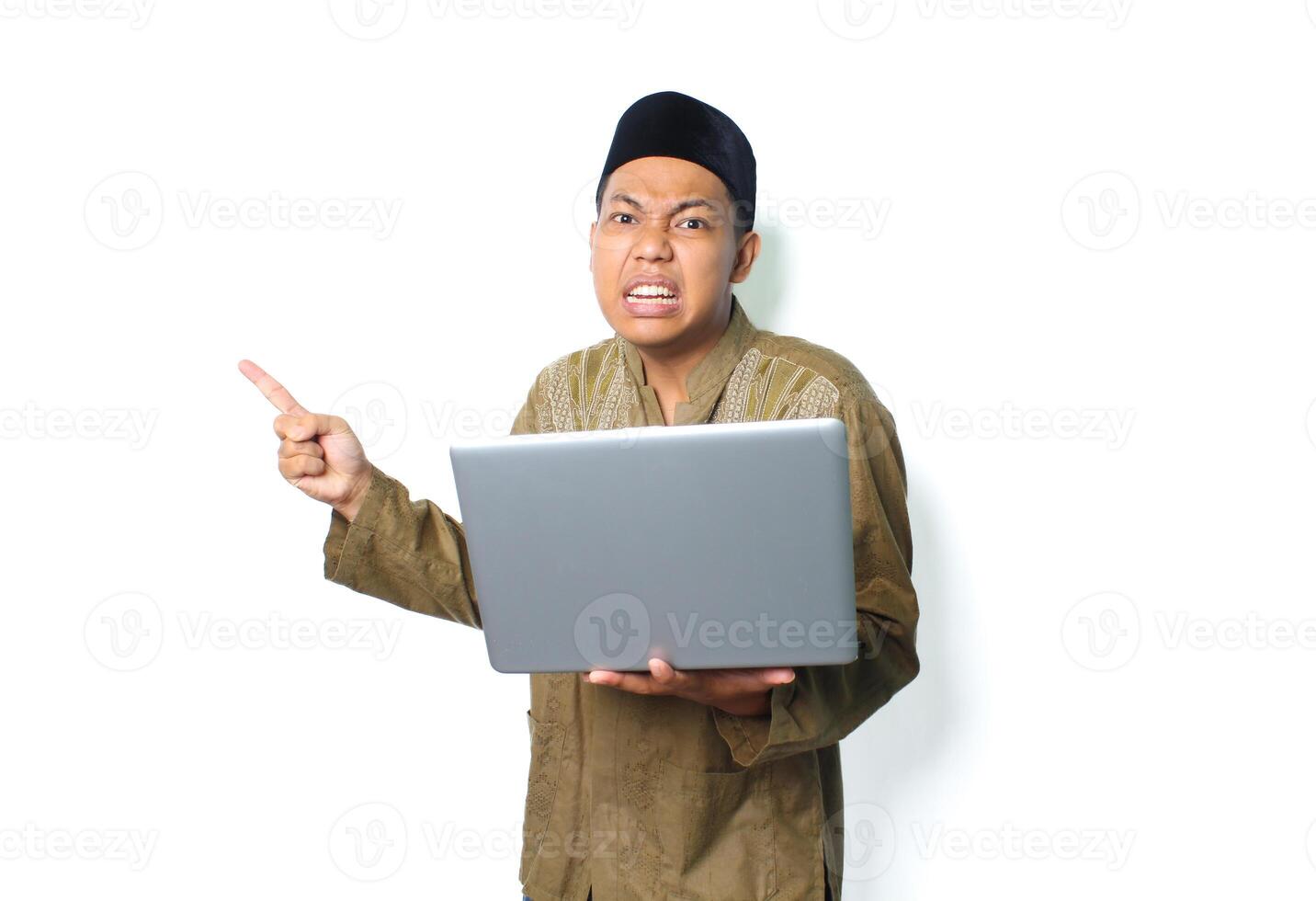 emotional asian muslim man pointing to beside with holding laptop wearing islamic clothes isolated on white background photo