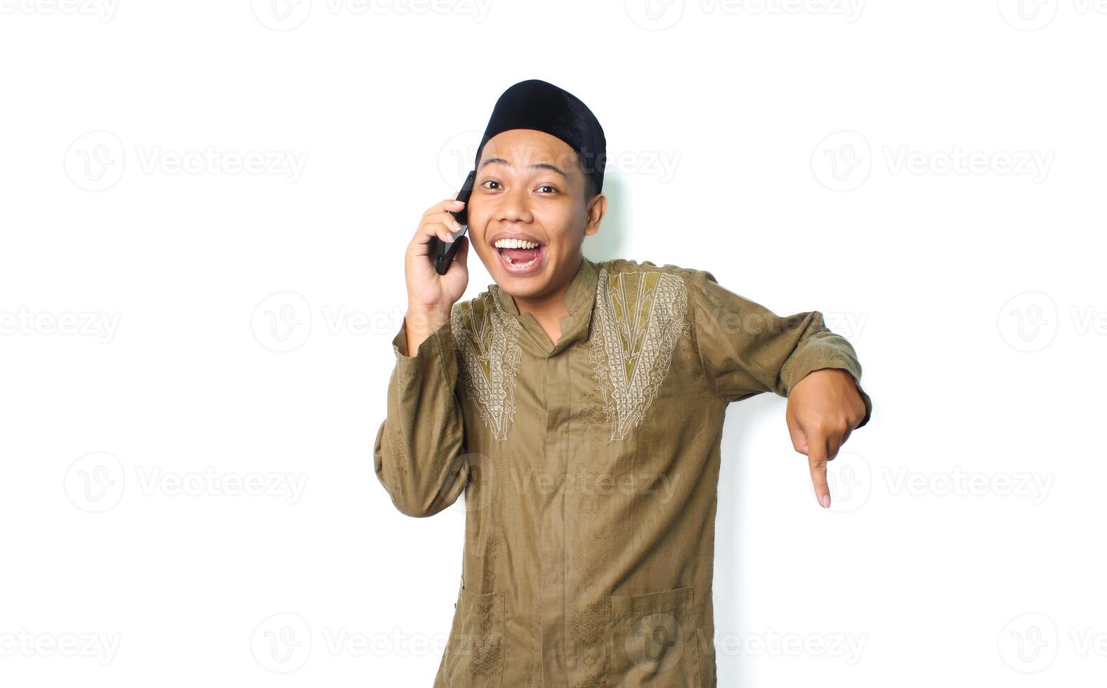 excited asian muslim male pointing down while talking using mobile phone isolated on white background photo