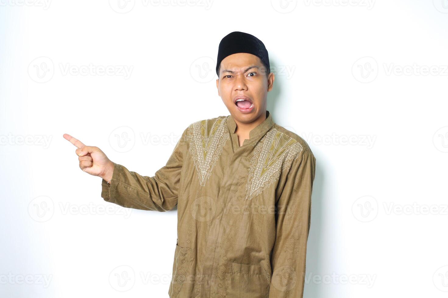 confused asian muslim man pointing to beside and looking at camera wearing koko clothes isolated on white background photo