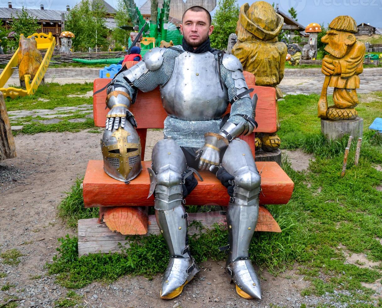 Knight in armor among wooden idols on a wooden bench. Knightly armor and weapon photo