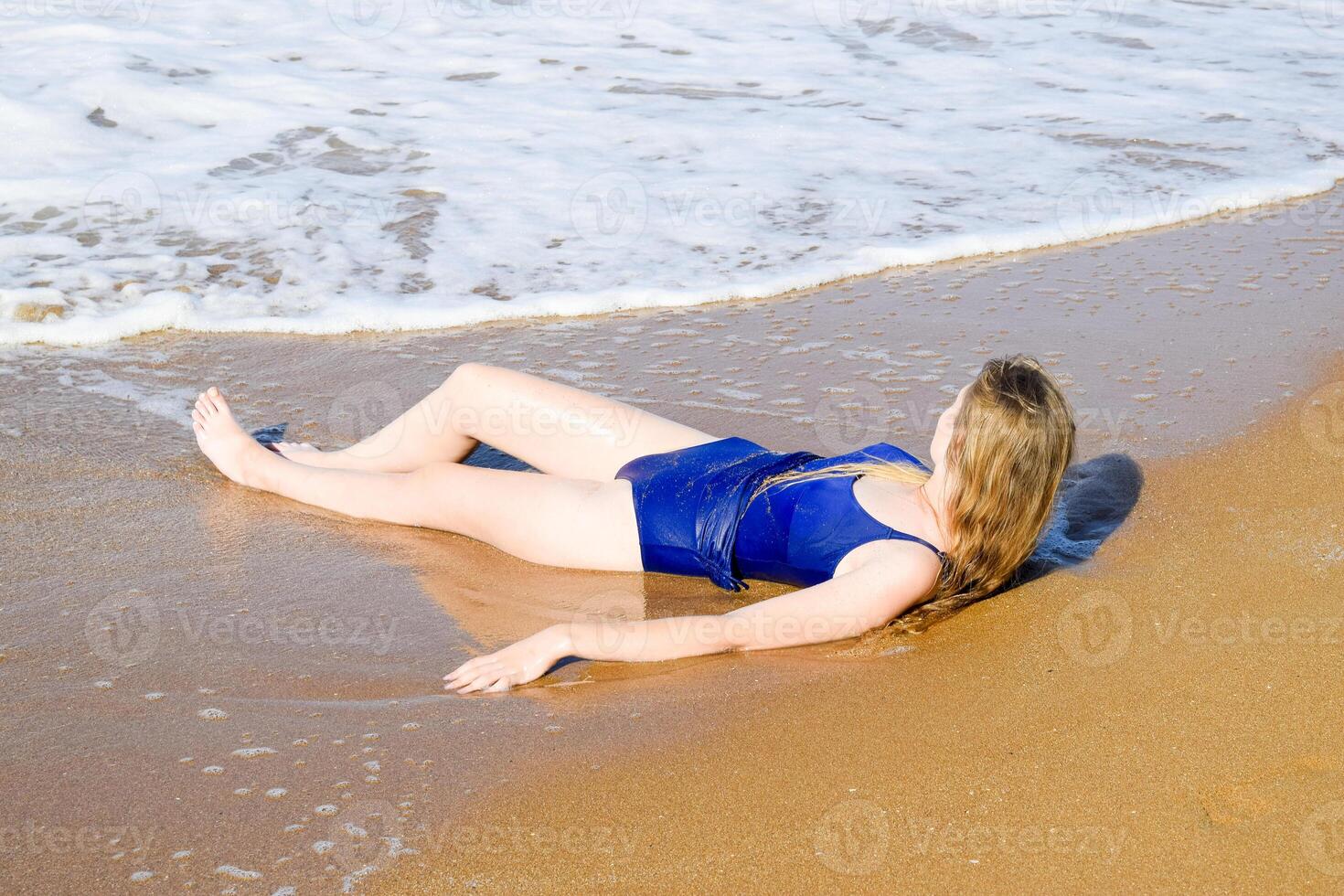 Girl in a blue bathing suit lies on the seashore and relaxes. Waves caress the girl's body photo