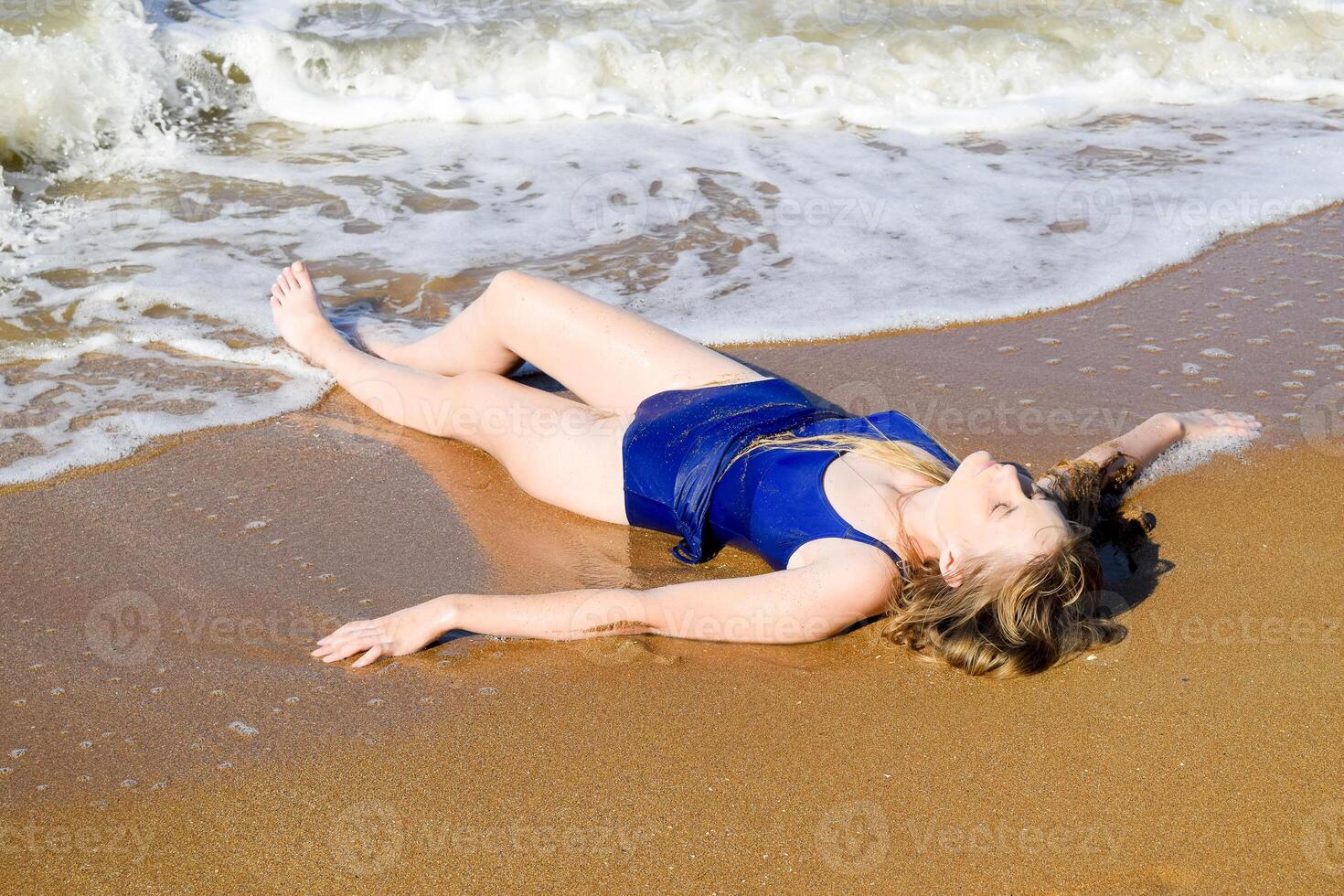 niña en un azul baños traje mentiras en el costa y relaja olas caricia el muchachas cuerpo foto