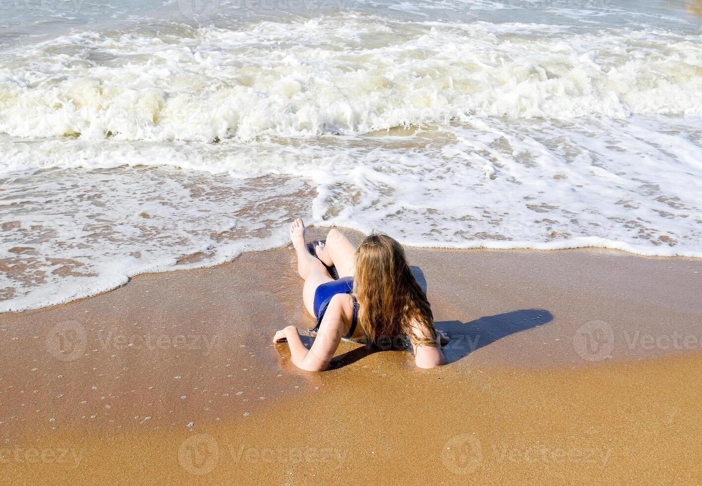 niña en un azul baños traje mentiras en el costa y relaja olas caricia el muchachas cuerpo foto