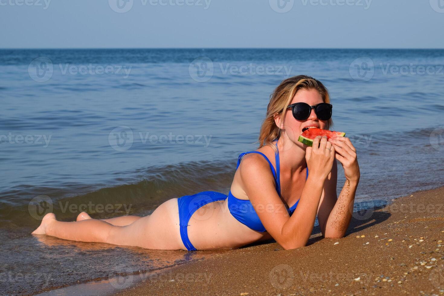 The blonde in sunglasses lies on the beach near the waves and eats a watermelon. A juicy watermelon in the hands of a woman. photo