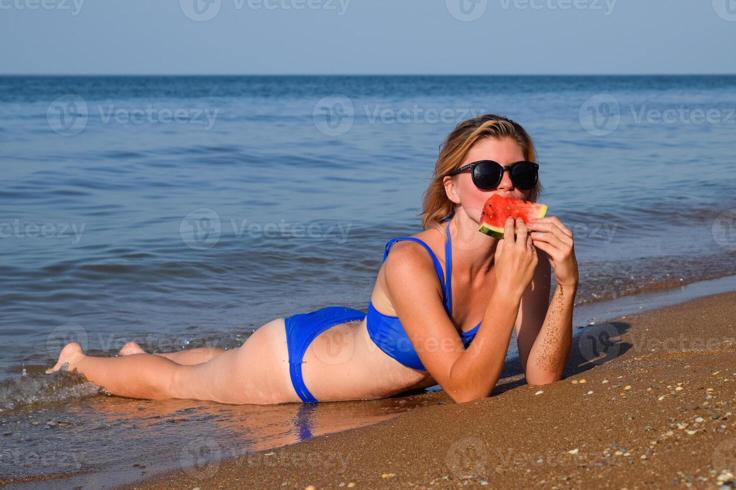 The blonde in sunglasses lies on the beach near the waves and eats a watermelon. A juicy watermelon in the hands of a woman. photo