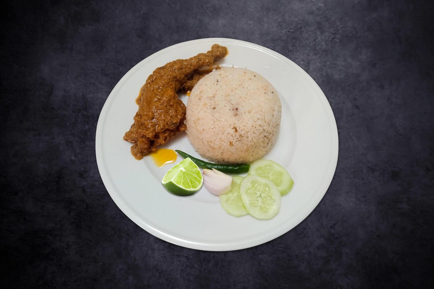 picante pollo asado con arroz servido con verde chile, cebolla, y Pepino en un blanco plato en negro antecedentes foto