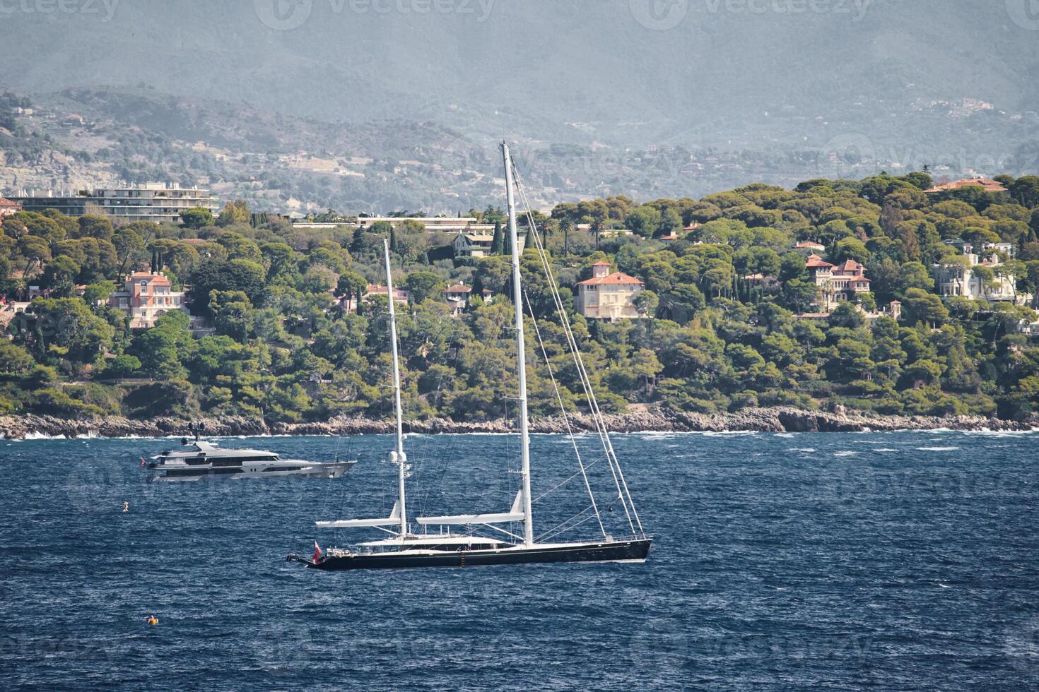 Mónaco, mega yate y enorme vela barco son en mar a soleado día, isla es cercano, enorme motor bote, riqueza vida de multimillonarios, montañas en antecedentes foto