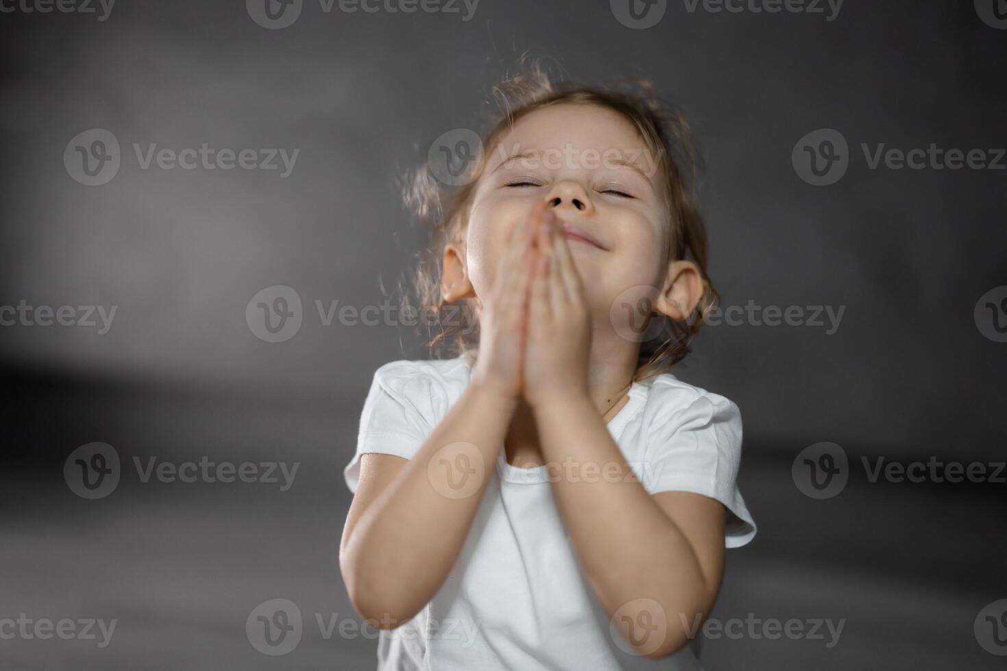 Tres años antiguo pequeño niña meditando en un loto actitud en un gris antecedentes en oscuro habitación foto