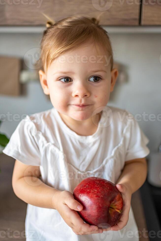 bebé niña rubia comiendo un manzana en el cocina, concepto de sano comida para niños foto