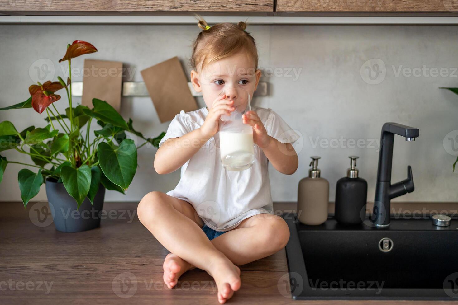 Happy baby girl sitting at the table in the kitchen and drinking milk photo