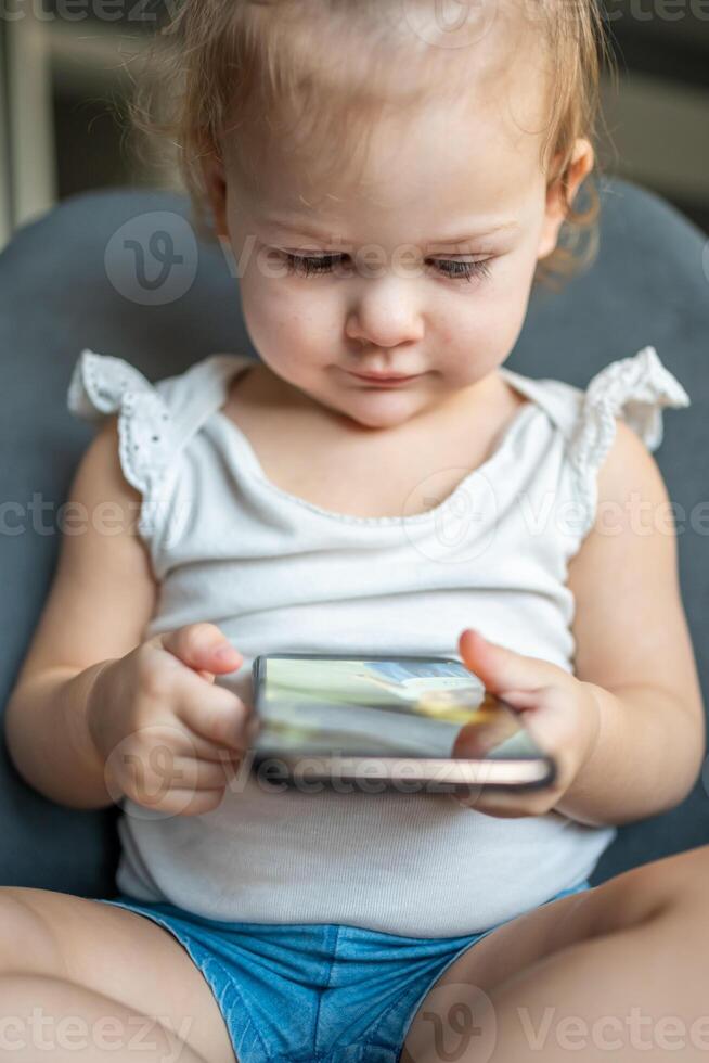 Smiling little girl is sitting on armchair and using smart phone photo