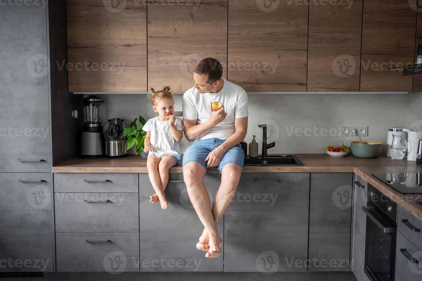 Cute little girl and her handsome dad are eating fruit in modern kitchen. Healthy eating. photo