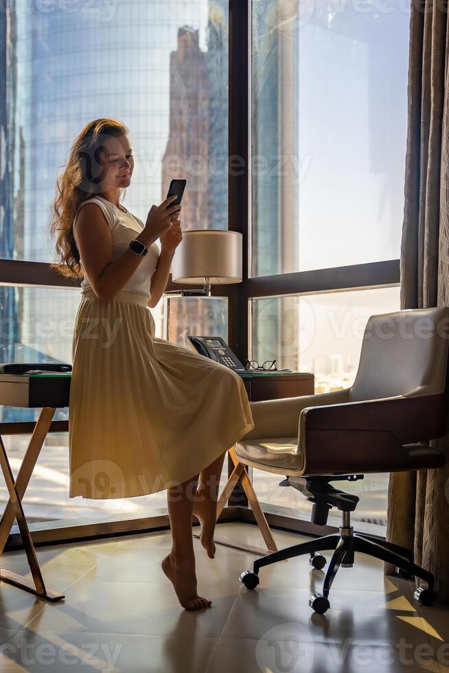 Stylish businesswoman freelancer working with smart phone and sitting on table, enjoying panoramic view in city background. Low key photo. High quality photo