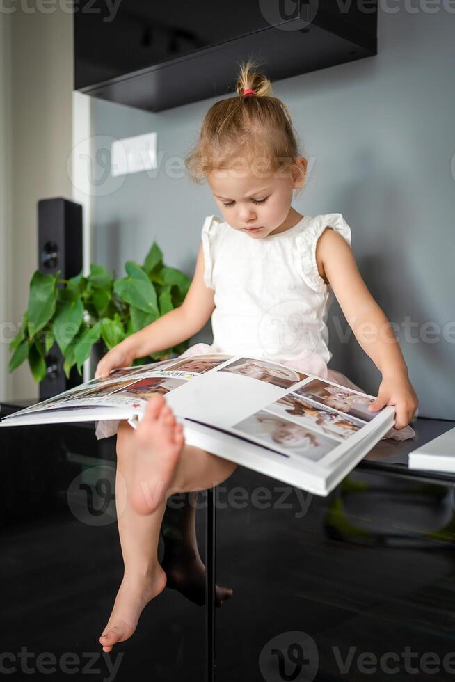 contento linda pequeño niña mirando a fotos en álbum. contento sonriente hija hojeada mediante impreso familia álbum, mirando a fotos.