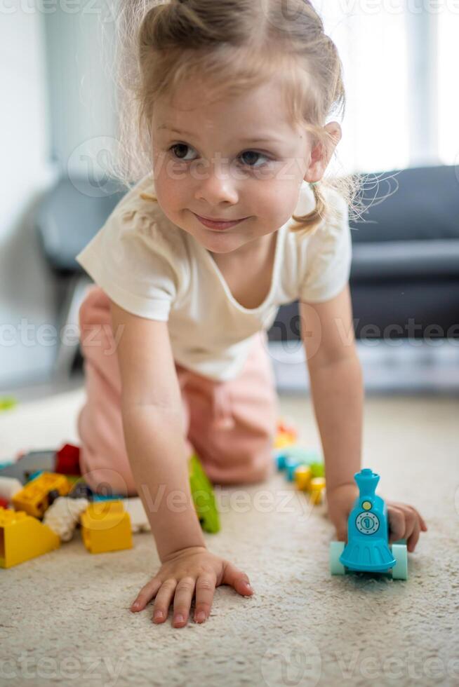 pequeño niña jugar con constructor juguete en piso en hogar, educativo juego, gasto ocio ocupaciones hora concepto foto