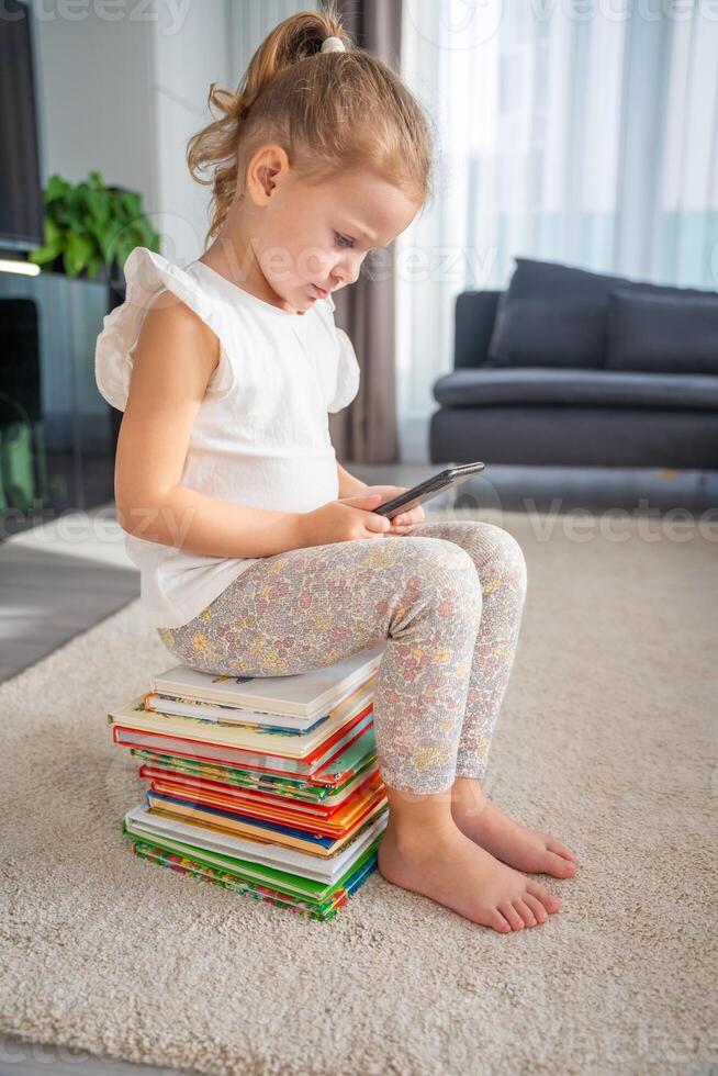 Little girl sits on a stack of children's fairy-tale books and plays the app on her smartphone photo