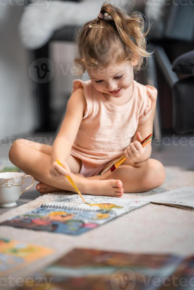 Little girl sits on the floor at home and draws with paints and brushes in a coloring book. Early childhood creativity and education. High quality photo