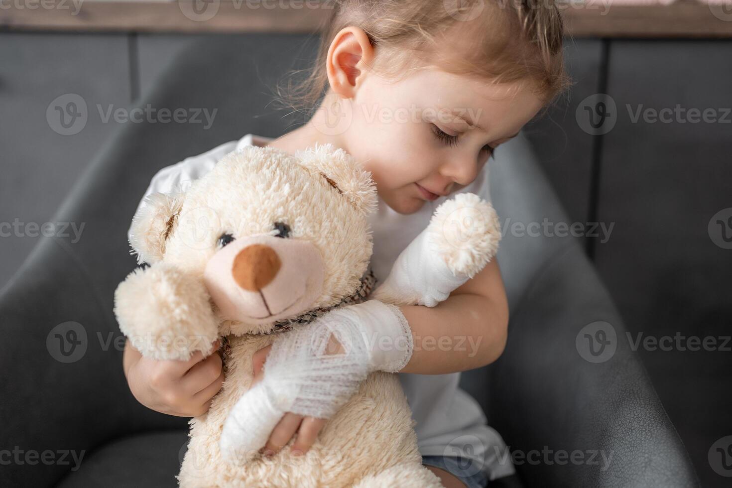 Little girl with broken finger holds teddy bear with a bandaged paw at the doctor's appointment in the hospital photo