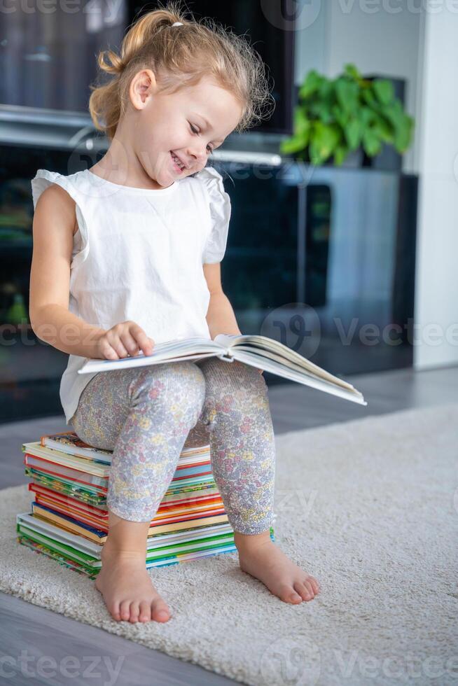 Smiling Little girl is sitting on stack of children's books and leafing through a book with fairy tales photo