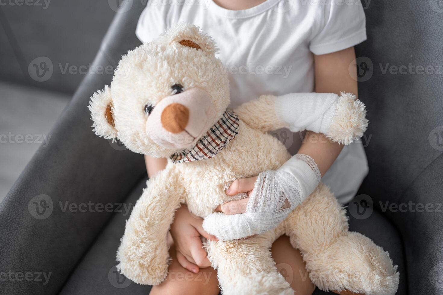 Little girl with broken finger holds teddy bear with a bandaged paw at the doctor's appointment in the hospital photo