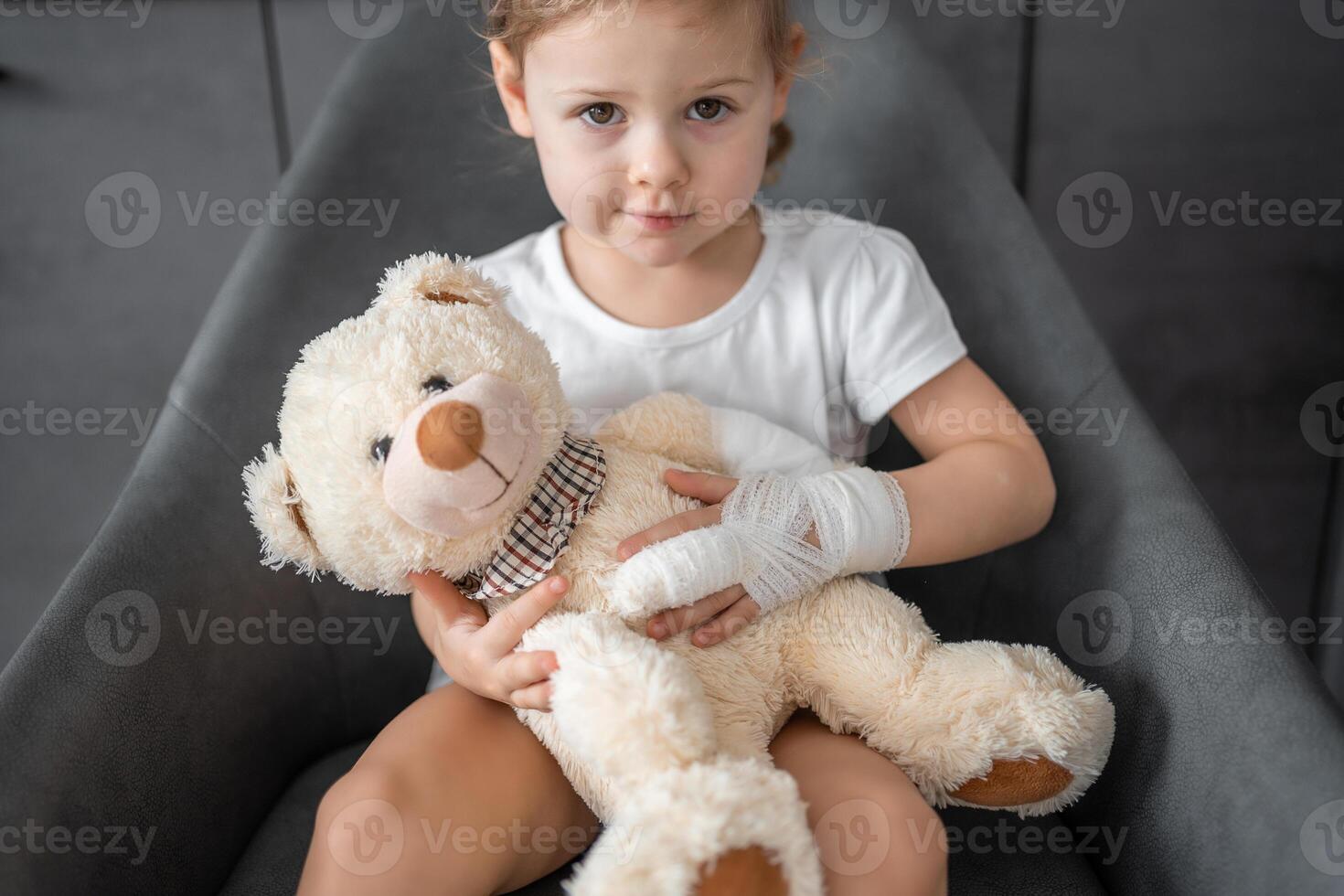 Little girl with broken finger holds teddy bear with a bandaged paw at the doctor's appointment in the hospital photo