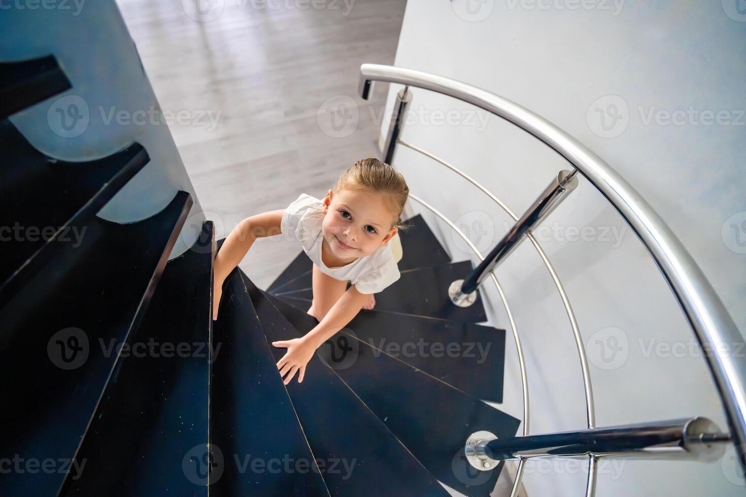 retrato de pequeño niño niña yendo arriba escalera a moderno hogar, niño alpinismo espiral escalera. peligroso situación a hogar. niño la seguridad concepto. foto