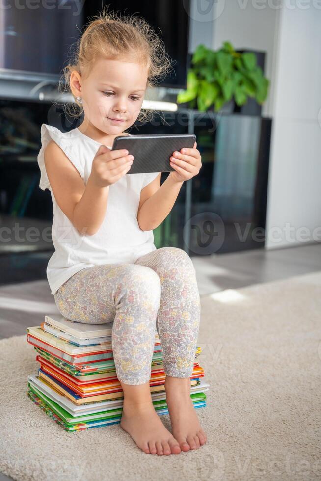 linda pequeño niña se sienta en un apilar de para niños cuento de hadas libros y relojes dibujos animados en su teléfono inteligente foto