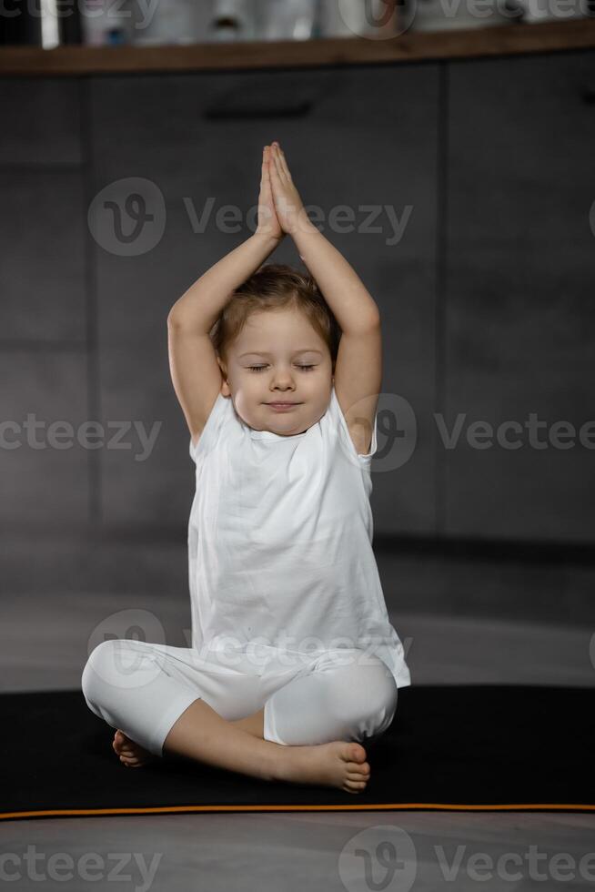 Three years old little girl meditating in a lotus pose on a gray background in dark room photo