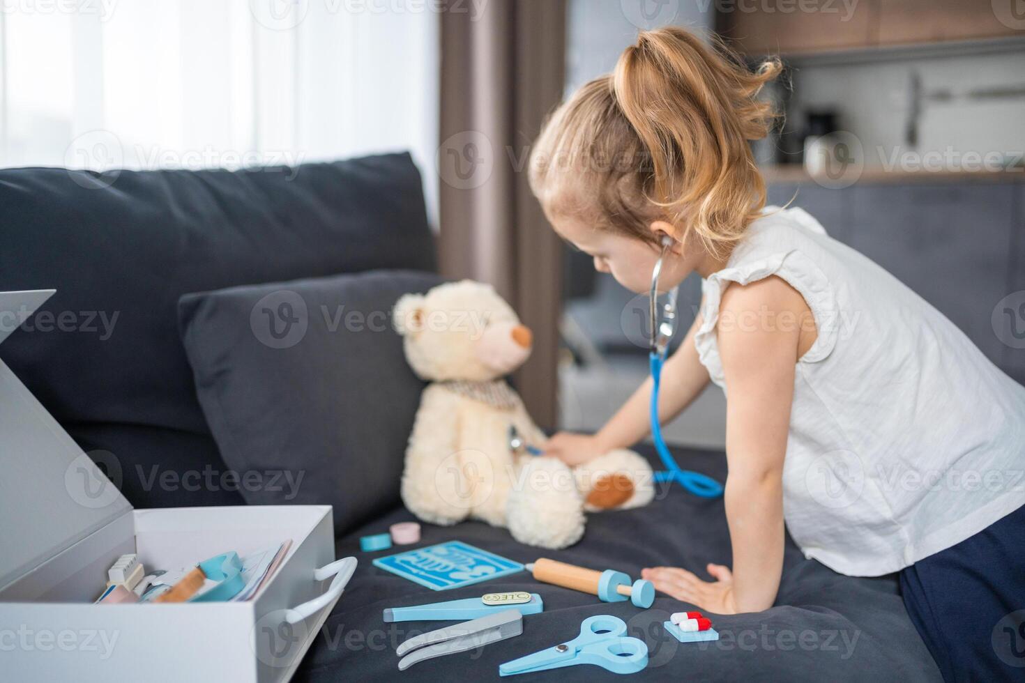 pequeño niña jugando médico con juguetes y osito de peluche oso en el sofá en vivo habitación a hogar foto