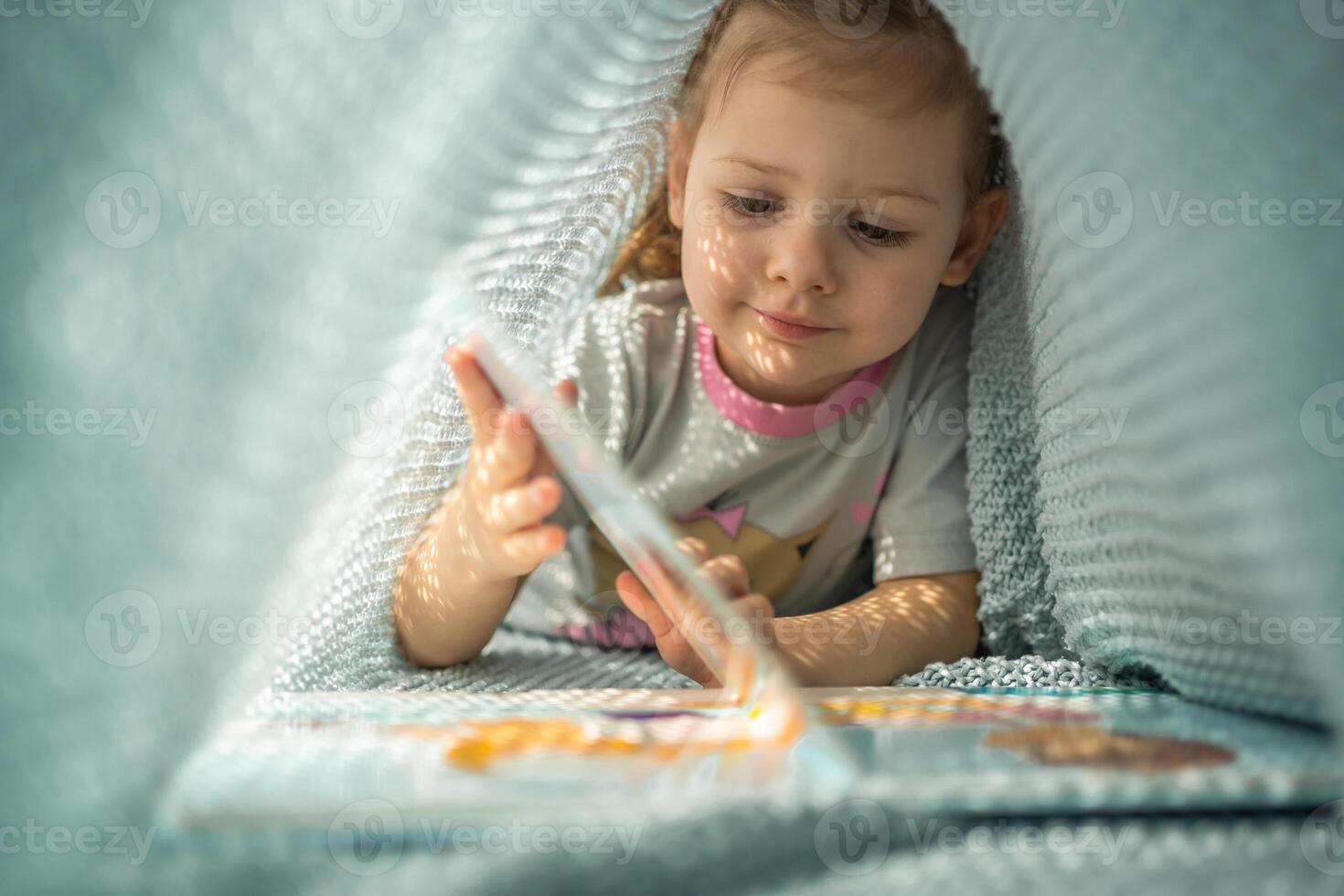 Little girl enjoying of review a book under blue knitted plaid in sunny morning photo