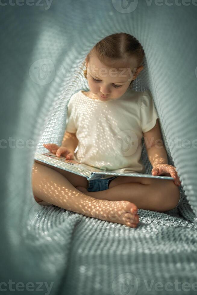 Little girl enjoying of review a book under blue knitted plaid in sunny morning photo